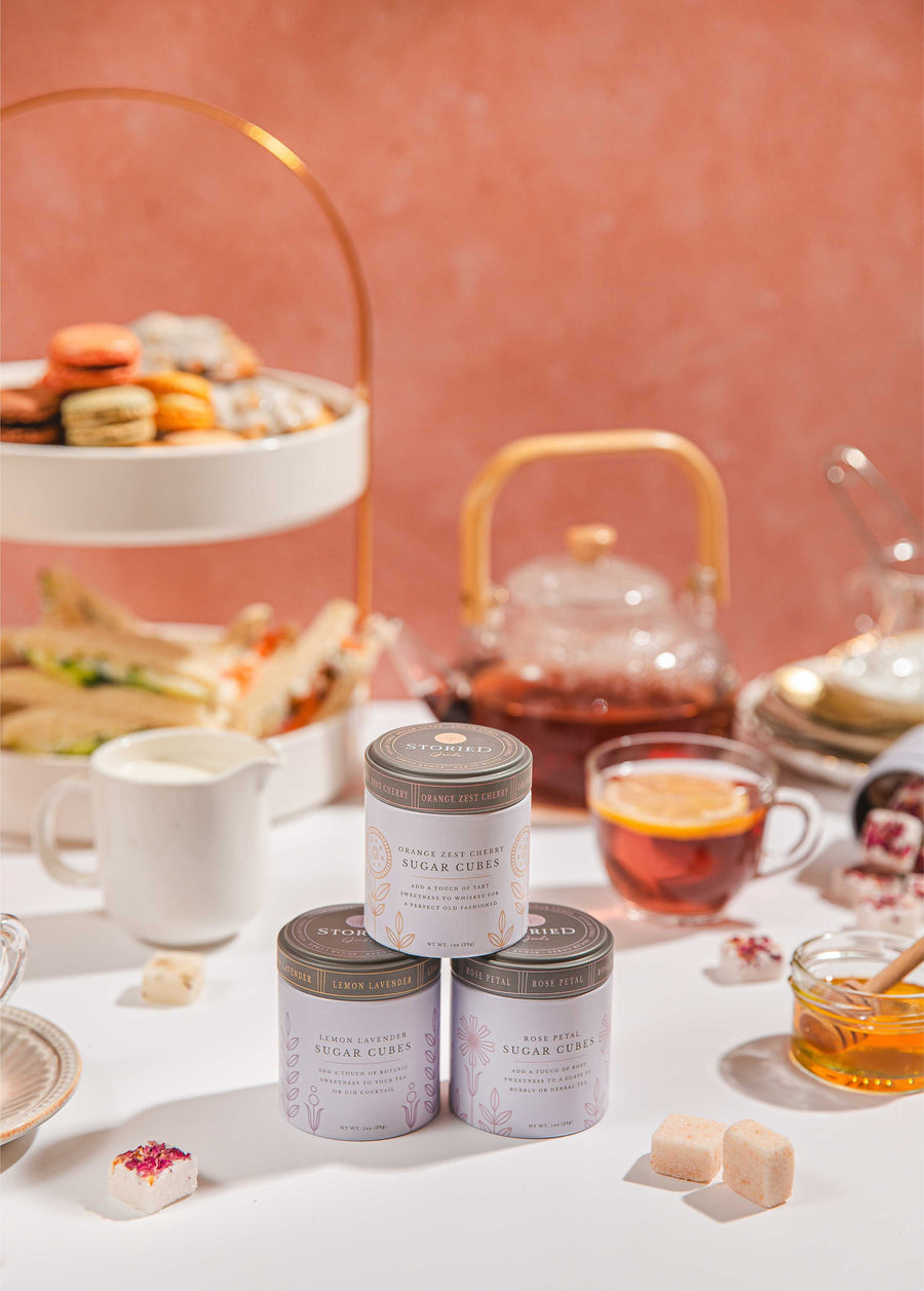 Tins of sugar cubes on a table surrounded by tea cups and a tea pot with a tray of sandwiches and biscuits